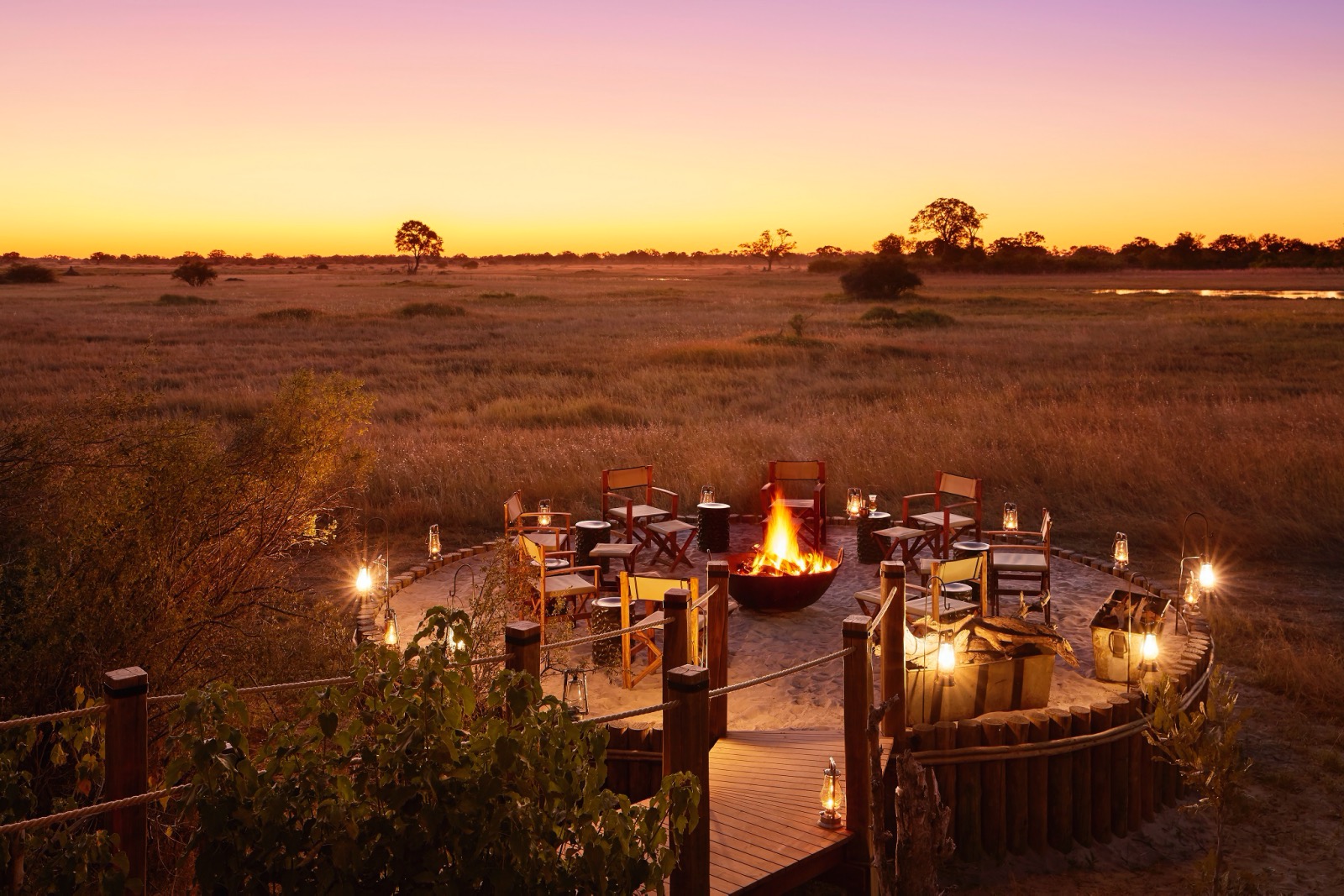 A fire burns brightly in the centre of an outdoor dining area overlooking the Botswana savanna and sunset